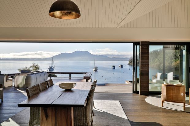 An open-plan waterfront dining and living area with expansive sliding glass doors framing a calm bay, moored boats, and mountainous surroundings. The interior features a long dark timber dining table with woven dining chairs, a low sculptural bowl, timber flooring, a white vertically panelled ceiling, a large dark pendant light, and a relaxed seating area opening onto a timber deck. The design has a warm, refined coastal character with strong indoor-outdoor connection.