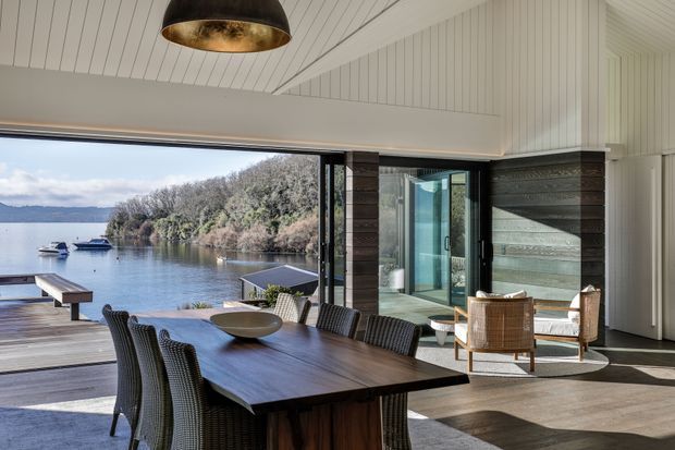 A refined open-plan dining and living space opening directly onto a timber deck beside a calm waterfront. The room features a long dark timber dining table with woven chairs, a sculptural pendant light, white painted vertical wall and ceiling boards, dark timber-clad structural piers, large sliding glass openings, and a seating area with woven armchairs. The view extends to the water, boats, and tree-covered shoreline.