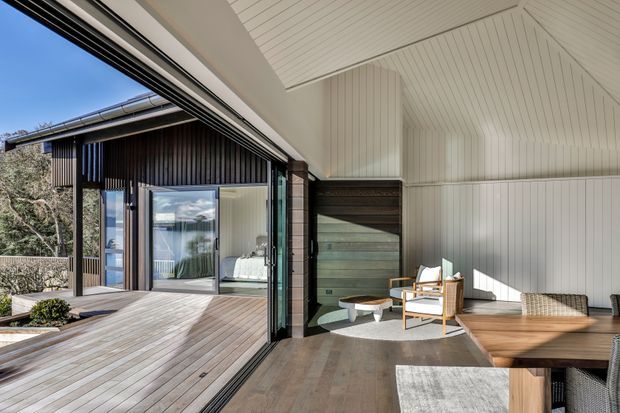 A bright contemporary open-plan living area opening onto a generous timber deck through large sliding glass doors. The interior features white vertical wall and ceiling panelling, a high sloping ceiling, dark timber wall cladding, built-in timber seating, and a light wood dining table. Natural light and views through the glazing create a strong indoor-outdoor connection.