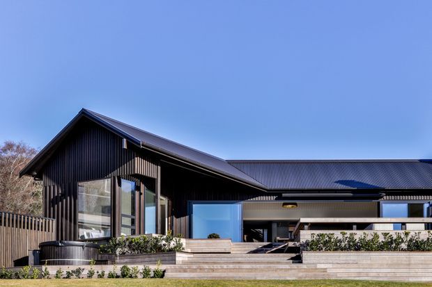 A contemporary single-storey house exterior with dark vertically ribbed cladding, intersecting pitched and low-sloping roof forms, large glazed openings, and broad timber-toned terraces and steps. The facade opens toward a landscaped outdoor living area with raised planters and a spa or plunge pool visible at the left.
