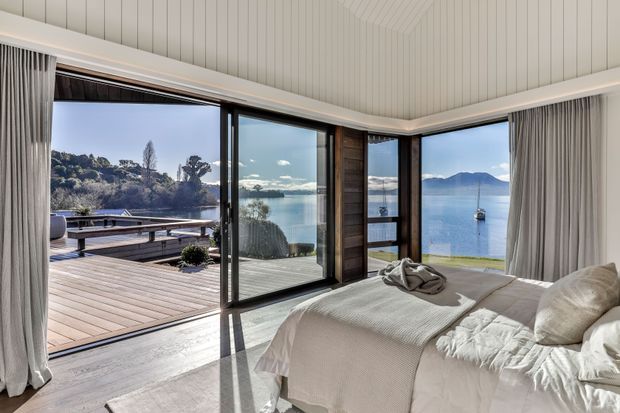 A bright coastal bedroom with floor-to-ceiling corner glazing opening onto a timber deck and expansive waterfront views. The room features a low-profile bed with soft off-white bedding, full-height curtains, a pale vertically panelled ceiling, dark timber window and door framing, and light timber flooring. A calm bay, moored boat, and wooded shoreline are visible outside.