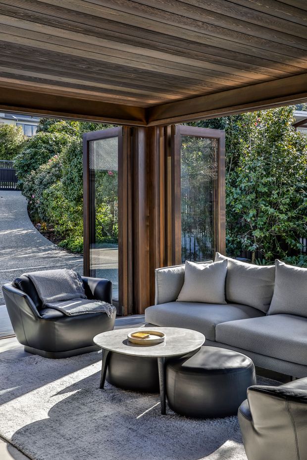 A refined indoor-outdoor living space with a grey sectional sofa, black leather lounge chairs and ottomans, and a round marble coffee table arranged on a large rug. Dark timber framing and timber-lined ceiling surround extensive glazed openings, looking onto a leafy garden. Bright natural light creates a sophisticated, comfortable atmosphere.