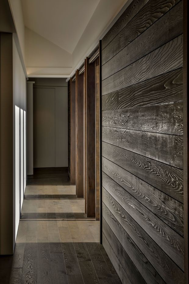 A narrow, softly lit interior hallway featuring a dramatic full-height wall of horizontally arranged dark, heavily grained timber boards. Matching dark timber-framed doorways and cabinetry line the corridor, while large-format grey-brown rectangular floor tiles and restrained grey walls create a moody, refined atmosphere. Linear daylight enters from the left, emphasizing the texture of the timber feature wall.