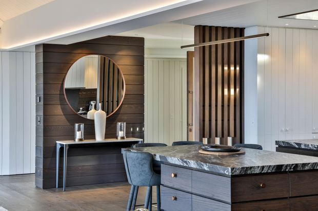 A refined open-plan kitchen and dining area with dark timber wall panelling, a large round mirror, slim console, marble-look island benchtop, dark cabinetry, and upholstered dining chairs. White vertical wall panelling and concealed linear lighting create a bright, contemporary interior with warm timber contrast.