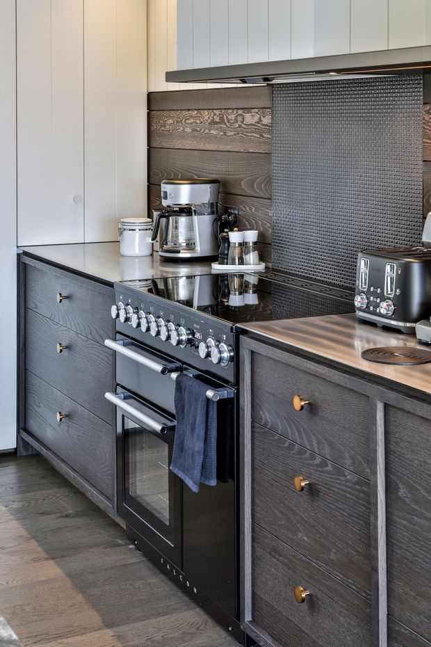 A compact, sophisticated kitchen featuring dark textured timber cabinetry, a black reflective benchtop and integrated rangehood, a freestanding black oven with metallic controls, and a dark textured splashback. White upper cabinetry provides contrast, while a coffee machine, toaster, and other small appliances sit on the counter.