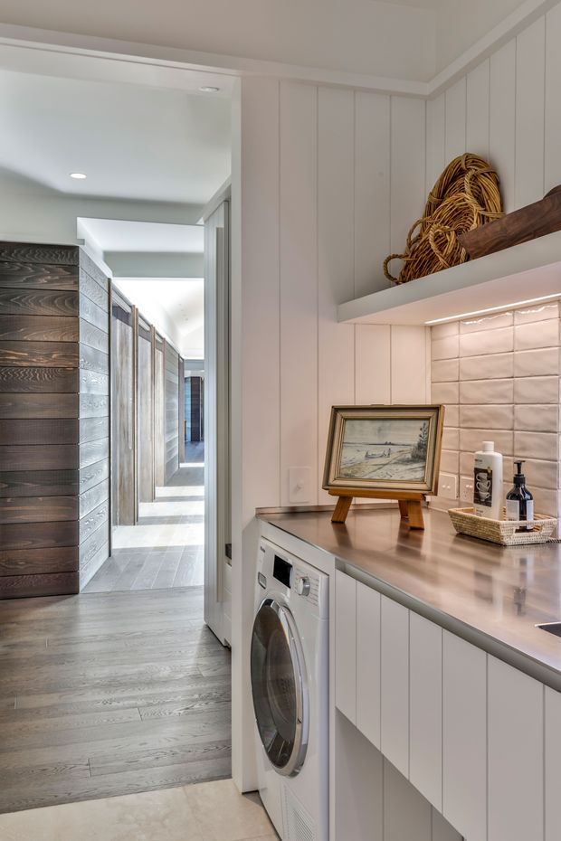 A bright, narrow laundry room with a front-loading washing machine integrated beneath a long grey benchtop. White vertical wall panelling, white cabinetry, a softly illuminated tiled splashback, open display shelving, and a basket with decorative objects create a clean coastal-style utility space. The laundry opens onto a light-filled hallway with timber wall cladding and pale flooring.