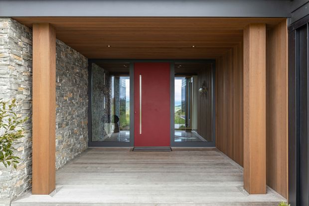 A contemporary covered entry porch framed by substantial warm timber posts and a timber-lined ceiling. The entry features a bold red pivot door with a narrow vertical metal handle, flanked by full-height glazing. Rough stacked-stone walls and grey timber-look flooring create a strong natural-material palette, while landscape is visible through the glazing.