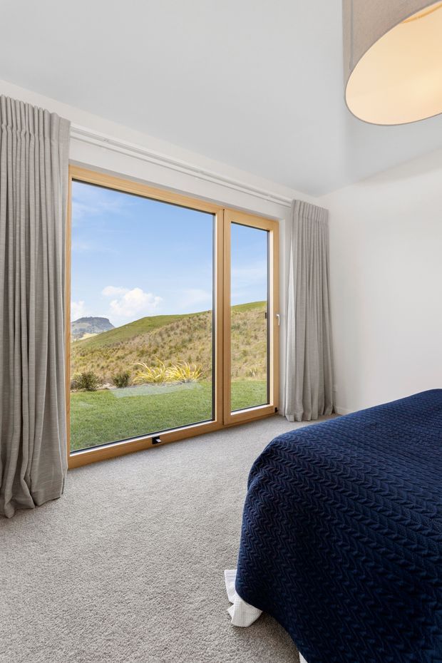 Bright, uncluttered bedroom with a large timber-framed glazed door or window overlooking a grassy hillside. The room has pale walls, light grey carpet, full-length grey curtains, and a bed dressed in a dark navy textured coverlet. A simple contemporary pendant light is partially visible overhead.