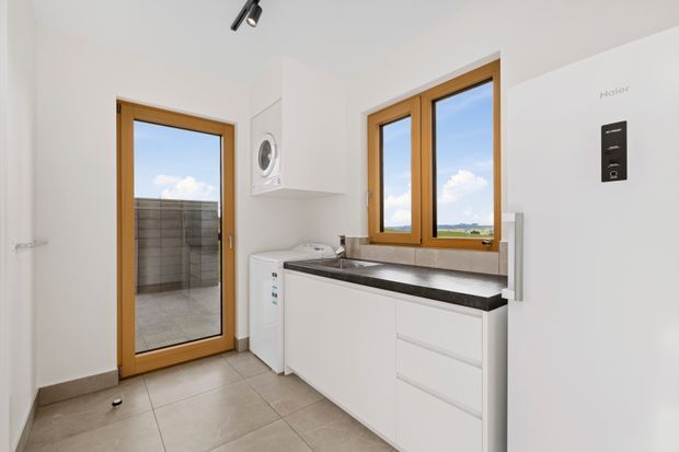 Bright contemporary laundry room with white built-in cabinetry, a dark stone-look benchtop, front-loading washing appliances, wall-mounted dryer, tiled floor, and warm timber-framed windows and external door. The room opens to a tiled outdoor terrace or balcony, with rural landscape visible through the glazing.