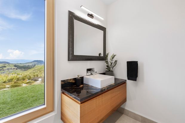 Bright contemporary bathroom with a floating timber vanity, white vessel basin, dark stone benchtop, large framed mirror, wall-mounted light, and black towel. A large window provides expansive views of a green rural landscape.