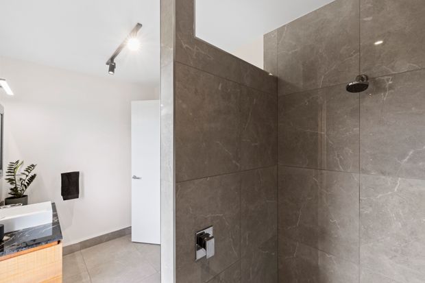 Contemporary bathroom with a walk-in tiled shower occupying the foreground. Large-format grey-brown stone-look tiles line the shower walls, with chrome fixtures and a clear glass partition. A white vanity with timber detailing, wall-mounted toilet, freestanding plant, and bright track lighting are visible beyond.