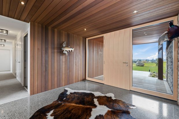 Contemporary entryway featuring extensive warm timber wall and ceiling panelling, a large timber pivot door with adjacent glazing, polished grey concrete flooring, and a cowhide rug. The glazed opening provides views toward a covered outdoor deck and open rural landscape. A white-carpeted hallway and recessed lighting are visible to the left.