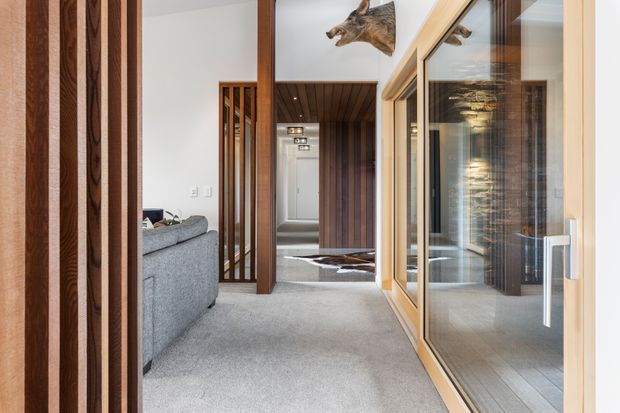 Interior view of a refined residential hallway or landing with grey carpet, warm timber wall and ceiling lining, vertical timber screens, and extensive glazed sliding doors. A living area is visible to the left, while the corridor leads toward a raised or stepped space with a wall-mounted taxidermy-style animal head and a dramatic illuminated stone feature wall reflected in the glazing.