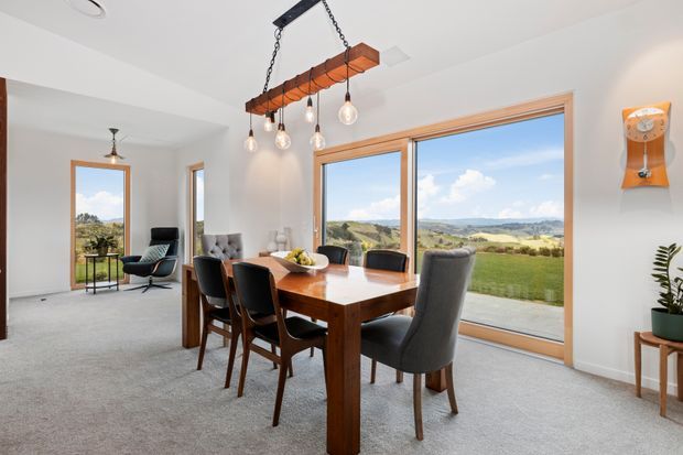 Bright dining area with a large solid timber dining table and upholstered chairs, positioned beside expansive timber-framed glazing overlooking a rural landscape. The room also includes a small sitting area, pendant lighting suspended from a timber beam, light carpet, white walls, and mid-century-inspired furnishings.
