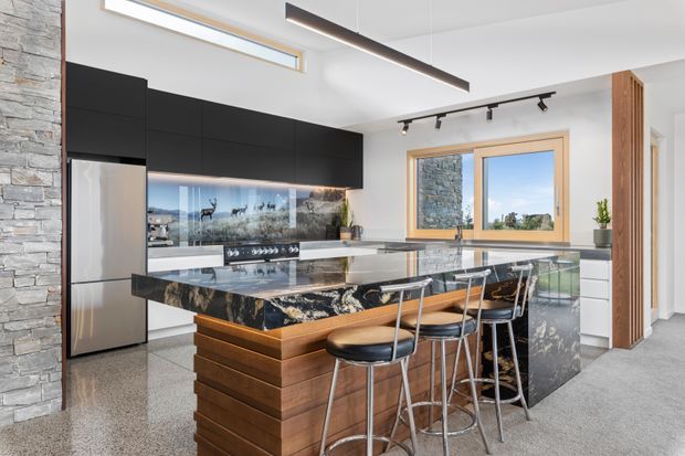 A contemporary kitchen with a large marble-look island featuring a waterfall edge and integrated seating for four. Matte black overhead cabinetry, stainless-steel appliances, reflective splashback, timber island detailing, grey carpet, stone-clad structural walls, timber-framed windows, and linear and track lighting create a refined, high-contrast interior.
