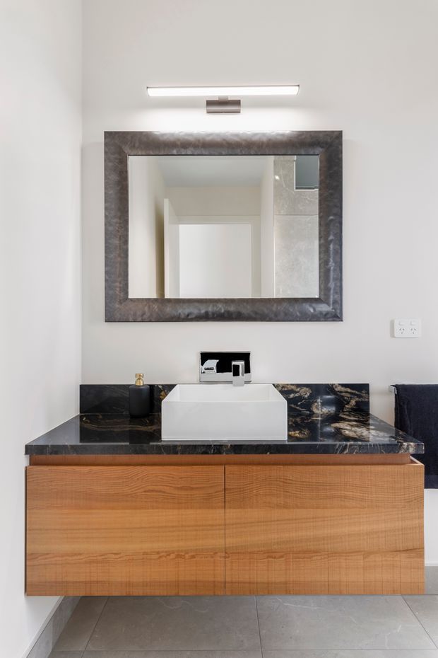 A contemporary bathroom vanity with a wall-mounted timber drawer cabinet, white rectangular vessel basin, chrome wall-mounted tap, and dramatic dark veined stone benchtop and splashback. A large dark-framed mirror and integrated horizontal light sit above the vanity, with pale grey tiled flooring and white walls.