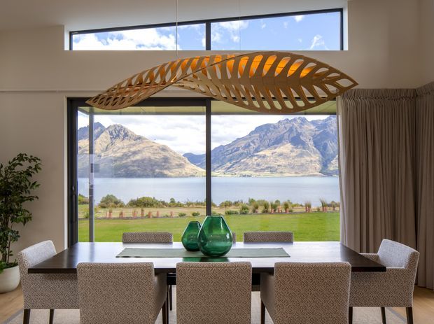 A refined dining room with a long timber dining table and upholstered dining chairs, framed by expansive floor-to-ceiling glazing overlooking a lake and mountainous landscape. A sculptural suspended timber pendant forms the main feature above the table, with neutral curtains, a potted plant, and green glass vessels adding understated colour. The room has a high ceiling and a strong indoor–outdoor connection.