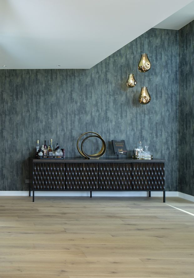A refined interior vignette featuring a dark textured feature wall, a long sculptural timber sideboard with patterned drawer fronts, and decorative objects including a circular metal sculpture, barware, and three hanging metallic pendant lights. Pale timber flooring and a white ceiling create contrast with the moody blue-grey wall finish.