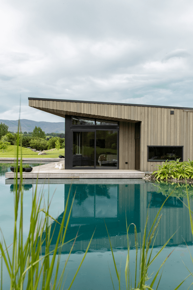 A modern single-storey pavilion or pool house beside a reflective swimming pool, clad in vertical pale timber boards with a projecting flat roof. Large dark-framed glazed doors open toward a light timber deck, while a horizontal window sits in the side wall. Reeds and planted greenery frame the pool, with a landscaped garden and distant hills beyond under an overcast sky.