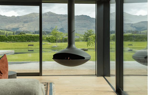 A contemporary interior with floor-to-ceiling glazing overlooking a green rural valley and mountains. The room features a suspended sculptural wood-burning fireplace with a dark opening, pale timber flooring, and a partially visible light upholstered sofa and patterned rug. Strong indoor-outdoor connection and expansive mountain views dominate the composition.