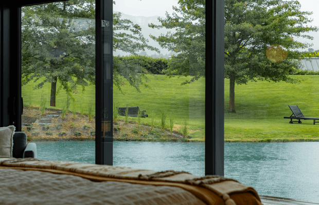 View from a bedroom through floor-to-ceiling dark-framed glazing, overlooking a tranquil turquoise water feature and expansive landscaped lawn with mature trees and outdoor loungers. The foreground includes the edge of a bed and upholstered seating, creating a calm indoor-outdoor connection.
