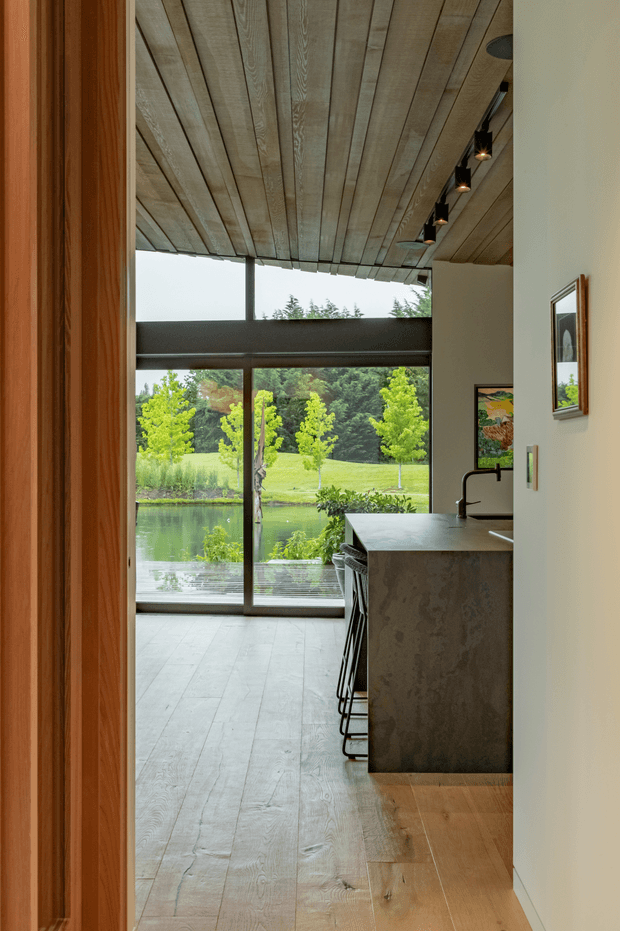 A contemporary open-plan kitchen and living area viewed through a timber-trimmed doorway. The space features a large dark stone island with bar seating, pale timber plank flooring, a matching exposed timber-plank ceiling, and extensive floor-to-ceiling glazing overlooking a landscaped garden and pond. Black track lighting, dark window framing, and white walls create a restrained, natural modern interior.