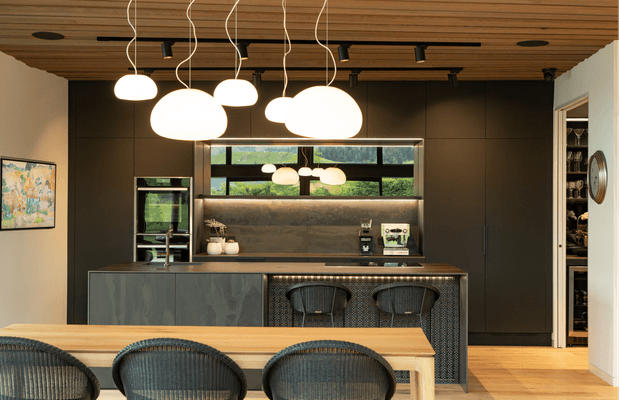 A refined contemporary kitchen and dining area with a dark, full-height kitchen wall, integrated appliances, illuminated stone backsplash, textured island/bar front, and timber dining table. Sculptural pendant lights hang from a linear black track beneath a warm timber slatted ceiling. A walk-in pantry or storage area is visible at right, while woven bar and dining chairs add natural texture.