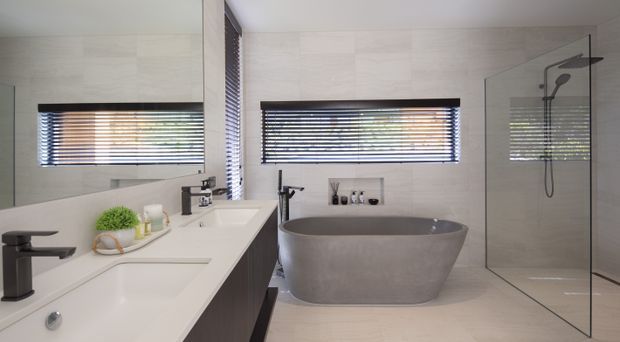 Contemporary bathroom featuring a freestanding oval grey bathtub, frameless glass walk-in shower, double white vanity with dark fixtures, recessed wall niche, and horizontal timber-look blinds. Large pale stone-look wall and floor tiles create a clean, neutral finish.