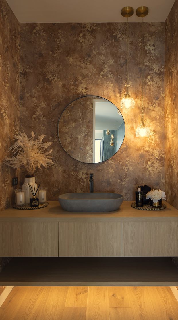 A richly styled bathroom vanity with a round black-framed mirror, sculptural vessel basin, wall-mounted timber-grain cabinetry, and warm pendant lighting. The walls feature dramatic brown textured decorative wallpaper or finish, complemented by wood flooring and bronze-toned accents. The reflection shows part of an adjacent light-filled space.