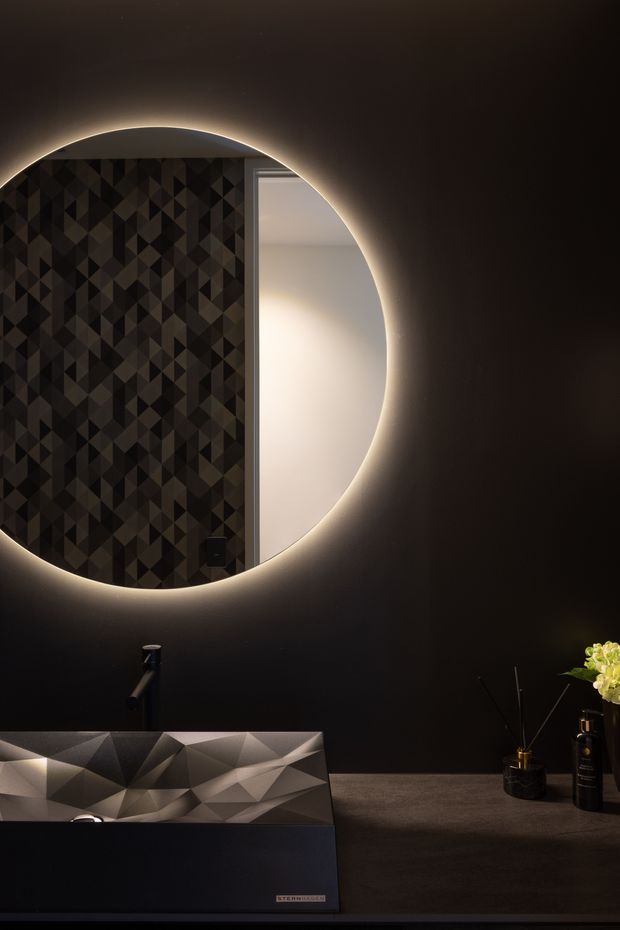 A dramatic, moody bathroom vanity scene featuring a large round backlit mirror, a dark geometric patterned wall panel, black wall surfaces, and a sculptural angular basin with a dark tap. Small bottles and a floral arrangement sit on the vanity, adding subtle detail to the luxurious dark interior.