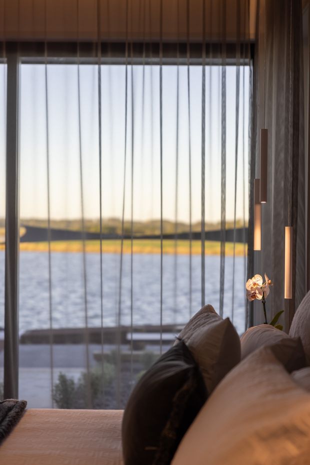 A close, atmospheric view from a furnished bedroom toward a large floor-to-ceiling glazed opening overlooking a body of water and distant shoreline. The foreground shows a bed with layered neutral pillows and bedding, dark textured wall finishes, a small flowering orchid, and slim vertical wall lights. Vertical glazing mullions or blinds create a strong linear rhythm, while warm low-angle light gives the room a calm, luxurious feel.
