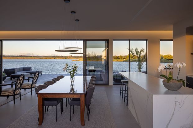 Contemporary open-plan kitchen, dining and living space overlooking a body of water through expansive floor-to-ceiling glazing. The room features a large white waterfall island with integrated seating, dark cabinetry, a formal timber dining table with upholstered chairs, sculptural pendant lighting, and polished pale flooring. Warm concealed ceiling lighting and sunset views create a refined, tranquil atmosphere.