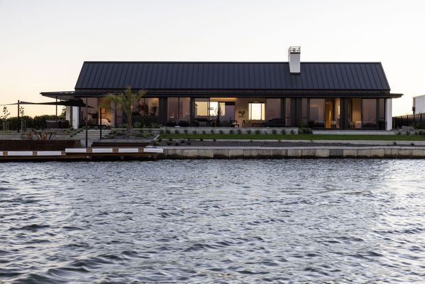 Long, low single-storey waterfront residence viewed across rippling water. The home has a dark standing-seam pitched roof with a central chimney, extensive floor-to-ceiling glazing, dark horizontal cladding, and a broad covered outdoor living area facing the water. A landscaped edge, deck and low retaining wall run along the shoreline.