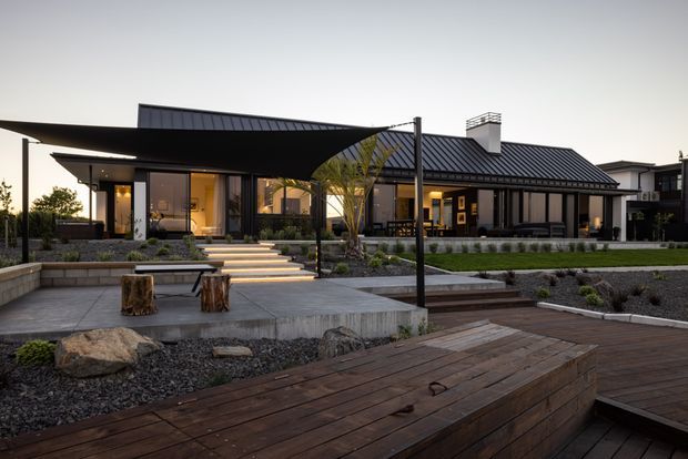A contemporary single-storey residence viewed from a landscaped outdoor deck and courtyard. The home has dark standing-seam pitched roofs, extensive floor-to-ceiling glazing, charcoal and timber-toned facade elements, and warm interior lighting. Stepped concrete paths, gravel garden beds, native-style planting, timber decking, and a small outdoor seating area create a refined modern landscape setting.