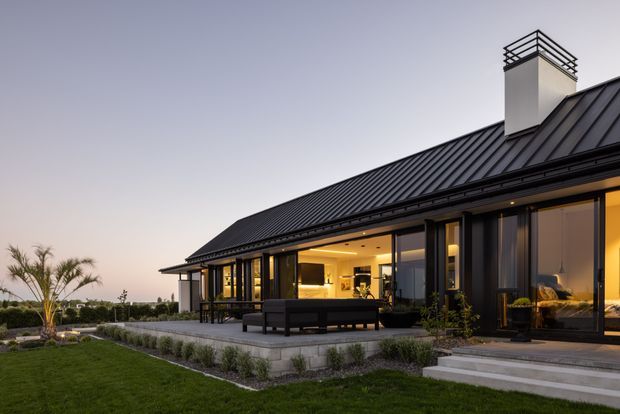 Exterior dusk view of a contemporary single-storey residence with a steep dark standing-seam metal roof, prominent chimney with black metal guard rail, extensive floor-to-ceiling glazing, and a covered outdoor terrace with built-in seating. The house opens onto a landscaped lawn and paved terrace, with warm interior lighting creating an inviting atmosphere.