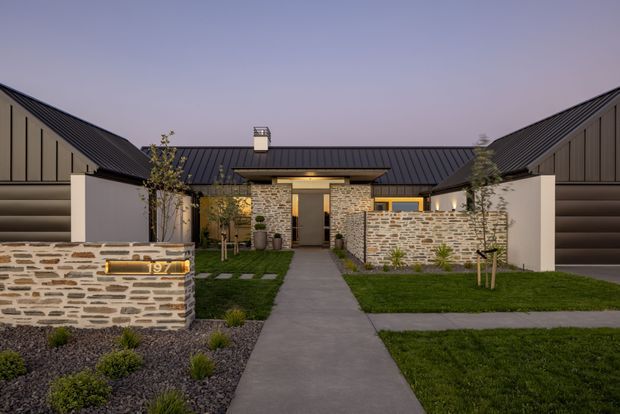 A landscaped front approach to a contemporary single-storey home, featuring dark standing-seam metal gabled roof forms, pale rendered walls, and extensive irregular natural stone masonry walls. A straight concrete path leads through neatly planted lawns and gravel garden beds to the recessed entry.