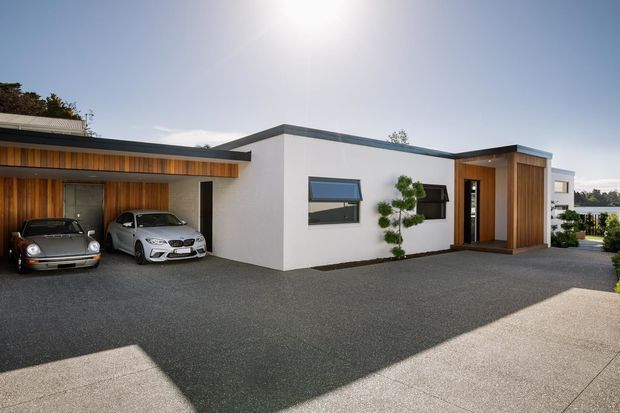 Exterior view of a low, single-storey contemporary home with crisp white rendered walls, dark flat rooflines, and warm vertical timber cladding around the covered carport and entry. Two cars are parked under the carport, while a paved forecourt and landscaped planting surround the building. A glimpse of water and neighbouring homes is visible in the background.