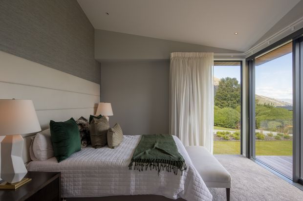 A refined bedroom interior with a neatly made bed, layered neutral and deep green cushions, a textured green throw, upholstered bench, bedside tables and lamps. Full-height glazed doors open to a landscaped outdoor setting and mountain views. The room has soft recessed lighting, pale carpet, textured wall covering and tailored curtains.