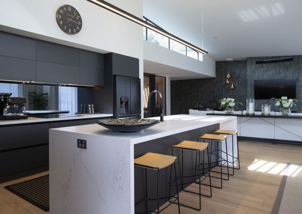 A spacious contemporary open-plan kitchen with a large white stone island, dark charcoal cabinetry, integrated appliances, timber flooring, and four slim bar stools. A double-height living area with a dark feature wall, wall-mounted television, clerestory glazing, and linear pendant lighting is visible beyond the kitchen.