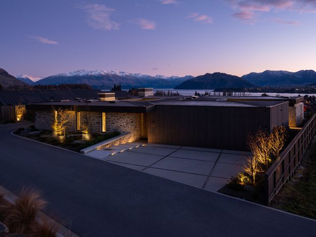 Twilight exterior view of a low-slung contemporary residence in a mountainous lakeside setting. The home features dark horizontal roofing, dark vertical cladding, warm illuminated stone walls, expansive concrete driveway paving, integrated landscape lighting, and a timber privacy fence. Snow-capped mountains and a lake are visible beyond the property.