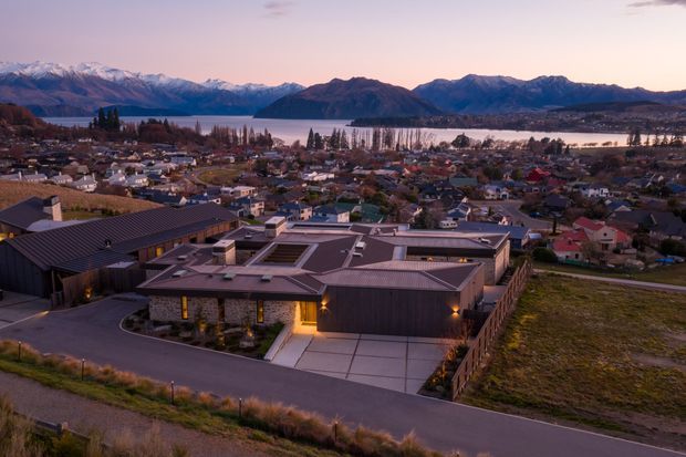 Aerial dusk view of a contemporary low-rise residential development on a sloping site, with flat-roofed buildings in the foreground and a dense town, lake, and mountain range beyond. The buildings feature dark metal-like roofs, timber-toned cladding, large glazed openings, landscaped entries, and paved driveways.