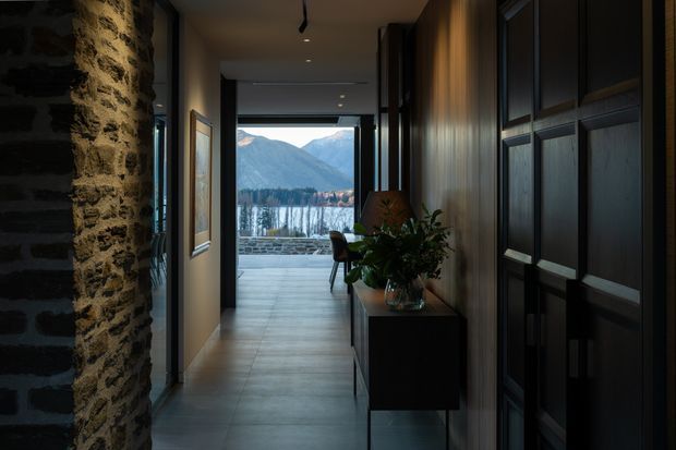 A dark, refined interior hallway or entry passage leading toward a large glazed opening and outdoor terrace, with dramatic mountain and lake views beyond. The corridor features a rough exposed stone wall on the left, dark timber or painted built-in cabinetry and doors on the right, smooth large-format grey floor tiles, warm wood wall panelling, recessed ceiling lights, and a slim console table with indoor greenery. The strong contrast between the shadowed hallway and bright alpine landscape creates a dramatic, luxurious atmosphere.