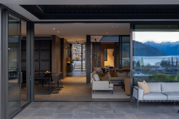 A refined contemporary home interior viewed from a covered outdoor terrace through wide-open glazed doors. The composition shows a connected dining area, living room and circulation spine, with dark timber and charcoal finishes, warm wood flooring, stone detailing, and expansive glazing framing a dramatic alpine lake and mountain landscape. A grey outdoor sofa occupies the foreground terrace, while the interior includes a dining table, upholstered seating, and a sculptural stone stair or feature wall.