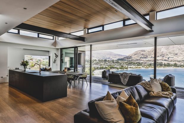 Open-plan contemporary kitchen and living area with a dark island, timber flooring, expansive floor-to-ceiling glazing, and a timber-lined ceiling with exposed steel beams. The room overlooks a lake and mountainous landscape, with a dining area and lounge seating arranged along the glazed facade.
