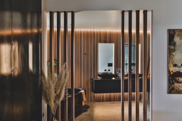 A refined contemporary bathroom viewed through a series of slim dark vertical screen battens. The room features warm timber slat wall panelling, a floating dark vanity with vessel basin, illuminated mirror, black fixtures, and warm concealed lighting. A freestanding dark bathtub is partially visible, while artwork and dried botanical decor appear at the edges of the frame.