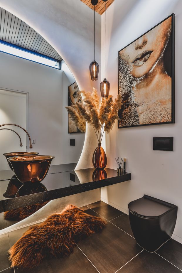 A dramatic, refined bathroom interior with dark tiled flooring, a wall-mounted black vanity shelf, sculptural amber glass vessel basins, a black toilet, large decorative portrait artwork, pendant lighting, and a tall vase of dried foliage. A curved ceiling detail with timber slatted lining and a high clerestory window adds architectural interest. The space uses reflective dark surfaces, warm amber accents, and strong contrast.