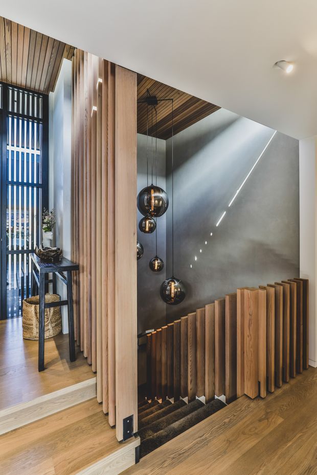 A contemporary interior stairway featuring warm timber flooring, vertical timber slat screening and balustrading, a dark textured wall, and a cluster of spherical pendant lights descending through the stair void. A tall glazed opening with vertical framing brings in daylight, while a narrow console table and woven baskets sit in the adjacent entry area.