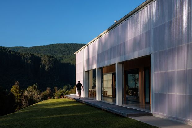 Exterior view of a contemporary, single-storey building set on a sloping landscaped site beside a forested mountainous backdrop. The long rectilinear facade features translucent corrugated panel cladding, large glazed openings, recessed timber-toned entry areas, and a narrow projecting deck or walkway. Strong late-day sunlight creates a calm, refined architectural composition.