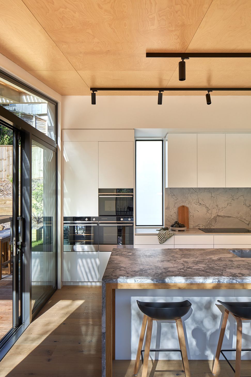 The kitchen is designed as the social heart of The Owl's Retreat, combining a refined material palette with everyday functionality. White cabinetry is paired with striking Super White granite benchtops, while a porcelain rear workbench and splash-back provide a durable and elegant workspace. Above, warm birch plywood ceilings soften the crisp finishes, adding natural texture and creating a welcoming atmosphere for cooking, gathering and entertaining.
