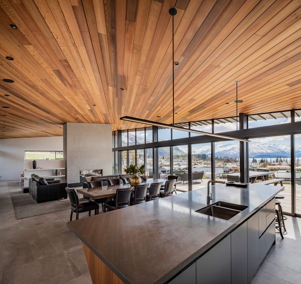 Open-plan contemporary kitchen, dining and living area with a large island, dark fixed cabinetry, expansive glazing and a richly detailed timber-lined ceiling. The space overlooks a town and mountain landscape, with a dining table and lounge area arranged beside the kitchen.